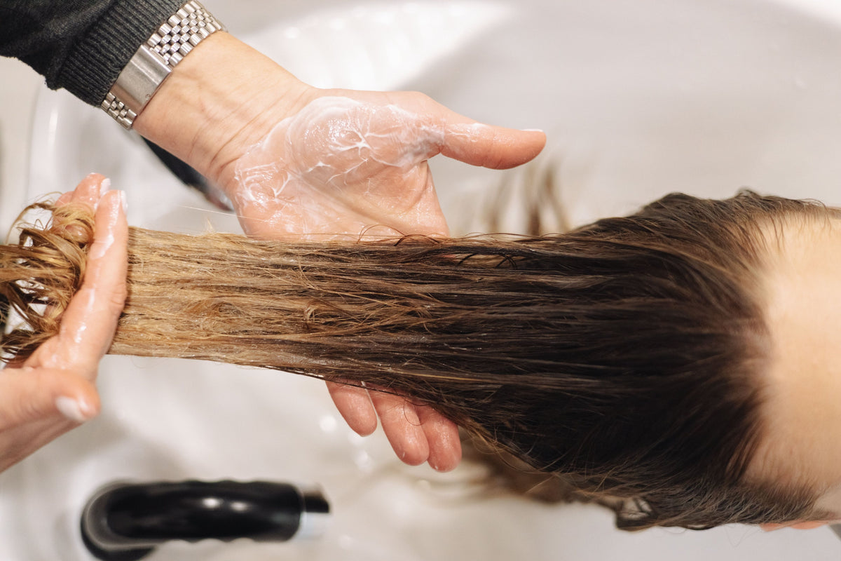 Manos aplicando tratamiento sobre cabello mojado en el lavacabezas